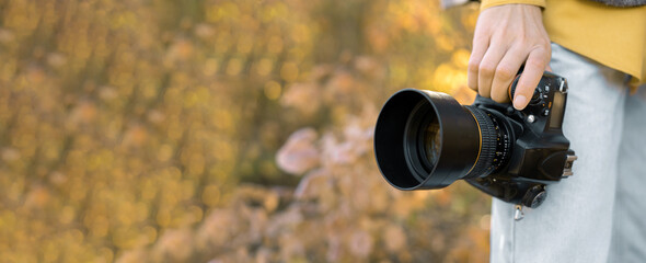 Close-up of a hand holding professional photo camera. A young lady stands in the autumn forest.