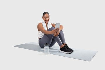 Black woman with a bright smile rests on a yoga mat, dressed in workout clothes, and enjoys a break...