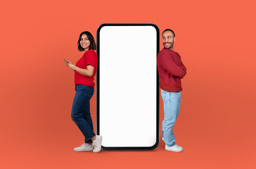 Multiethnic couple stand on either side of a giant, blank smartphone screen, both smiling and engaging with the empty display