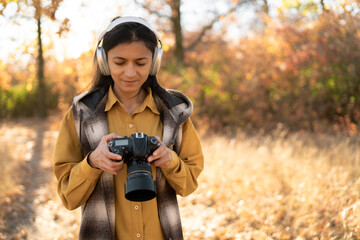 young beautiful woman photographer in headphones walks through the autumn forest in casual clothes. Holds a professional camera in his hands and sets it up.