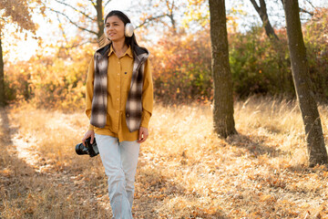 young beautiful woman photographer in headphones walks through the autumn forest in casual clothes. He holds a professional camera in his hands and looks for a beautiful landscape.