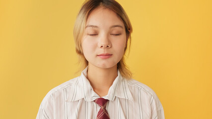Young woman dressed in shirt and tie stands with her eyes closed isolated on yellow background in...