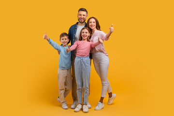 Happy family consisting of a mother, father, son, and daughter stands close together, all giving a thumbs up sign. They are posing against a vivid yellow backdrop