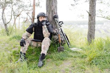 A man in a military uniform and a bulletproof vest sits in the forest near a metal detector and a...