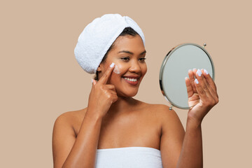 A smiling young woman with a towel wrapped around her head is gently applying cream to her face with her fingers, while holding a handheld mirror to see her reflection, against a soft beige backdrop.