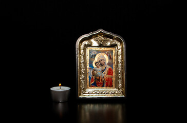 A lighted candle next to the icon of the Mother of God with the baby Jesus in her arms, on a black background. Decorative or ornamental image