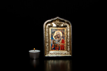 A lighted candle next to the icon of the Mother of God with the baby Jesus in her arms, on a black...