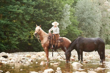 Cowgirl crossing the river riding a horse and leading a mule