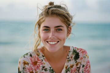 A close-up of a cheerful young woman with a floral top, smiling against a serene beach backdrop.