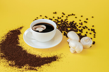 A white coffee cup and saucer stand on a yellow background. A slice of champignon mushroom floats in a cup, and coffee beans and ground coffee are scattered nearby and three champignons lie. mushroom 