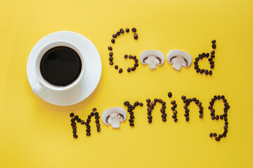 A white coffee cup and saucer stands on a yellow background. Next to it is a good morning phrase made from coffee beans and pieces of champignon mushrooms. mushroom coffee