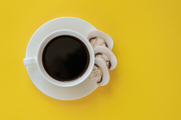 white coffee cup with saucer shot from above on a yellow background. There is coffee in the cup and slices of champignon mushrooms in the saucer. mushroom coffee