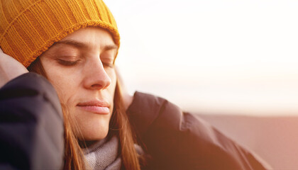 Portrait of a young woman with closed eyes in a yellow knitted hat on the beach near the sea in the evening at sunset in winter, close-up, soft selective focus
