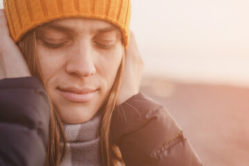 Portrait of a young woman with closed eyes in a yellow knitted hat on the beach near the sea in the evening at sunset in winter, close-up, soft selective focus