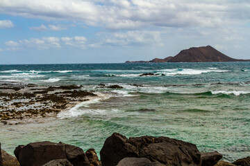 The rocky, volcanic coastline of the Atlantic Ocean near the port of Corralejo. Labos island in the...