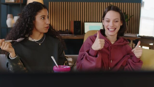 Medium shot of Caucasian girl with cerebral palsy speaking to Biracial female friend sitting on couch in front of TV with pint of ice cream