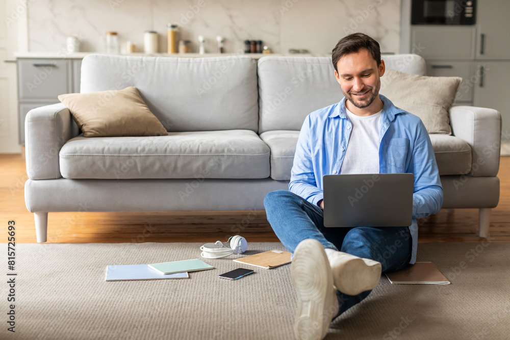 Aufkleber A man is seated on the floor with a laptop open in front of him. He appears focused on the screen, typing and interacting with the device. The room is dimly lit, and there are no other people present.