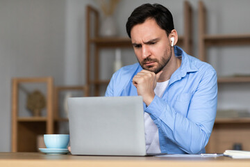 A concentrated man is seated at a desk in a well-lit room, looking intently at his laptop screen....