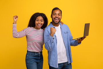 Black couple standing side by side against a yellow backdrop, both exuding excitement and joy. The...