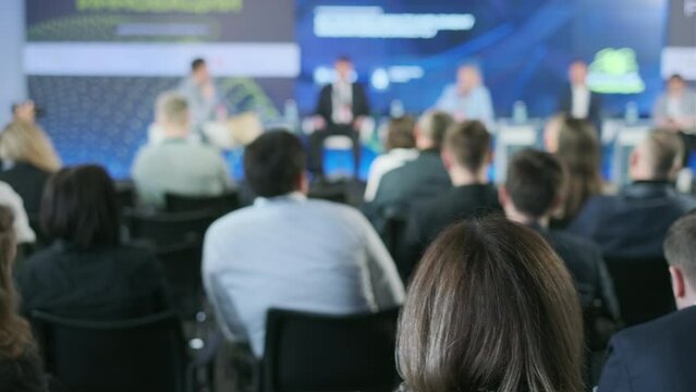 Blurred view of an engaged audience at a conference with speakers in the background, reflecting a professional setting and knowledge exchange.