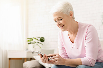 Elderly woman is seated on a couch indoors, engrossed in her cell phone. She appears focused as she...