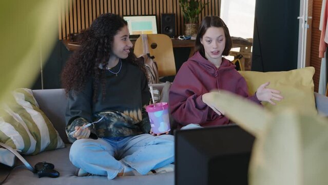 Medium shot of two multiethnic teenage girls sitting on couch in living room and chatting while watching film on TV with pint on ice cream on weekend night, one of girls having cerebral palsy