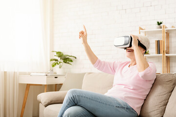 Elderly woman is seated on a couch, wearing a virtual reality headset, engaged in the virtual...