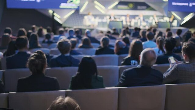 A room filled with professionals attentively listening to speakers on a stage at a business conference. Useful for illustrating corporate events, networking, and professional development.