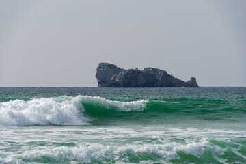 Une belle vague couleur émeraude se forme dans les eaux turquoises de la mer d'Iroise, avec en arrière-plan le majestueux rocher du Lion, créant une image captivante de la nature sauvage.