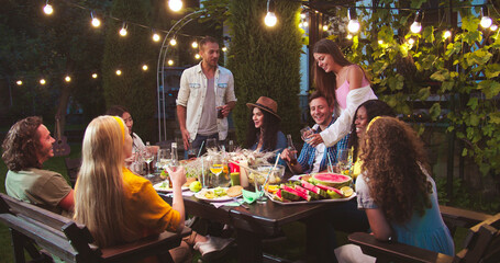 Happy multi-ethnic friends chat at garden in evening. Caucasian man gives high five to girl. African American girl sitting at table. Asian woman laughing. Friends raising and clinking glasses - Powered by Adobe
