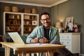 A portrait of a smiling male consultant holding a cup of coffee in front of a laptop in his home...