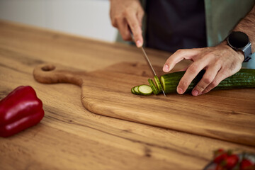 A close-up shot of man's hands cutting a fresh cucumber on a wooden board with a kitchen knife