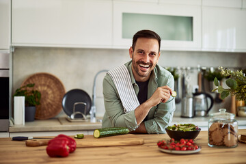 A portrait of a happy adult bearded man preparing a fresh salad and eating a cucumber as a snack...