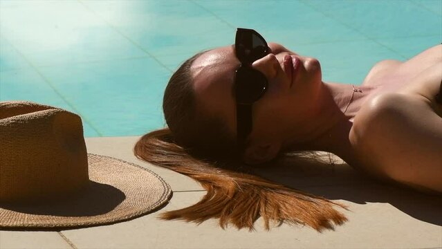 Beauty fashion model girl lying near pool and relaxing, sunbathing. Beautiful young woman portrait over pool background, sun tan concept. Slow motion. Vacation. 