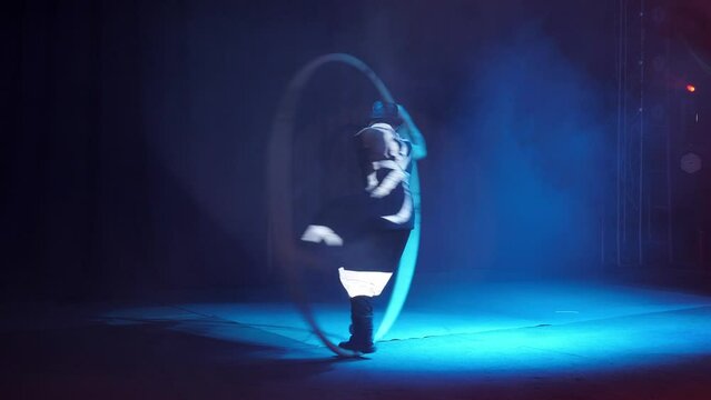 A muscular artist rotates on a circus gymnastic wheel in a circus arena, under the light of colorful spotlights and smoke. A gymnast is balancing on a large circus wheel. Concert lighting.
