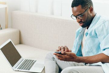 Smiling African American man working on laptop and talking on phone while sitting comfortably on a sofa in his modern apartment The freelancer enjoys the convenience of online shopping and