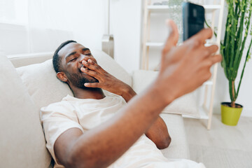 Happy African American man sitting on a black sofa, typing on his mobile phone The modern apartment...