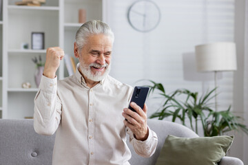 Excited senior man celebrating success while using smartphone at home