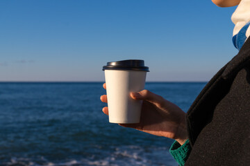 Close-up of a woman's hand holding a white paper disposable cup with coffee or tea against the background of the sea on a sunny day, copy space