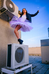 a ballerina in tutu, jacket and boots climbed on the air conditioner and poses against the sky