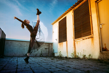 a ballerina in a black skirt and bodysuit does vertical splits on the roof of a building against...