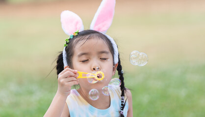Happy 6 year old Asian little girl with bunny ears blowing soap bubbles in park, having fun, sunny...