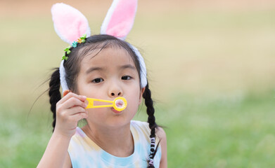 Happy 6 year old Asian little girl with bunny ears blowing soap bubbles in park, having fun, sunny day. Portrait of cute child playing outdoors in summer in nature