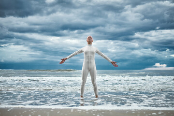 Happy hairless girl with alopecia in white futuristic suit stands with spread arms on beach bathed...