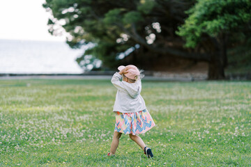 Little girl walks on a green lawn holding a cap on her head. Back view