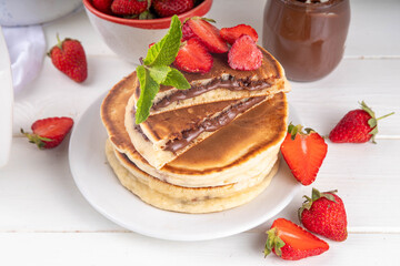 Stack of chocolate nutella filled pancakes. Sweet breakfast pancakes with chocolate sauce inside it, and fresh stawberries, on white kitchen table