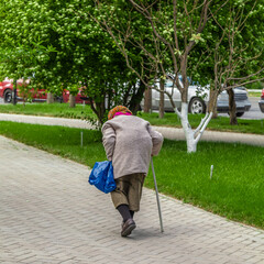 Old woman with walking stick. Pensioner. Woman with bag walks down road. Life in city.
