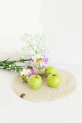 Purple and white carnation flowers with green apples lying on beige wicker table napkin. Handmade round tablecloth