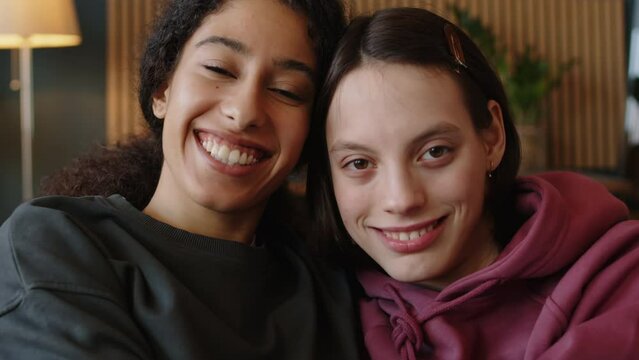 Tilt up portrait of Caucasian teen girl with cerebral palsy and her Biracial female best friend sitting on sofa embracing each other and smiling at camera