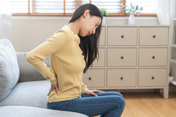 Pain body muscles stiff, suffer asian young woman painful with back pain, back ache sitting on sofa...